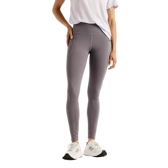 Der ses en person fra skuldrene og ned, iført en hvid kortærmet skjorte, hvide sportssko og et par New Balance Reflective Rc Heat Legging 27″ til kvinder i farven Neptune Grey. Leggingsene med høj talje har reflekterende detaljer og sidelommer. Baggrunden er ensfarvet hvid.
