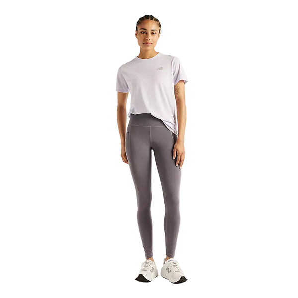 En person står med ansigtet vendt fremad iført en lyselilla T-shirt, hvide sportssko og New Balance Reflective RC Heat Legging 27″ til kvinder i farven Neptune Grey – leggings med NB HEAT-teknologi – foran en ensfarvet hvid baggrund, hvilket udgør et ideelt sæt tøj til løb i koldt vejr.