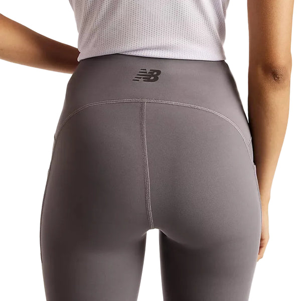 Der vises et billede af en person set bagfra fra livet og ned, iført New Balance Reflective Rc Heat Legging 27″ i farven Neptune Grey. Modellen med høj talje er udstyret med NB HEAT-teknologi og et synligt New Balance-logo på linningen, hvilket gør disse leggings ideelle til løb i koldt vejr. Personen har desuden en hvid, struktureret top på.