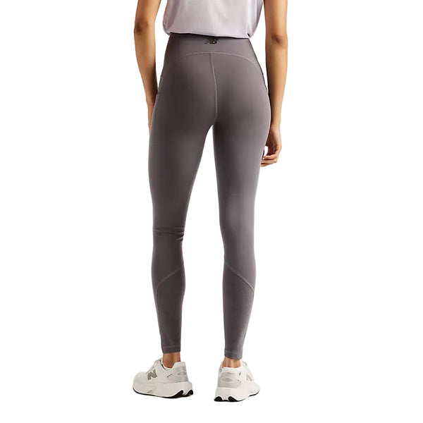 En person står med ryggen til, vist fra taljen og ned mod en ensfarvet hvid baggrund, iført New Balance Reflective Rc Heat Legging 27″ til kvinder i farven Neptune Grey med NB HEAT-teknologi, kombineret med en hvid top og hvide sneakers – perfekt til løb i koldt vejr.