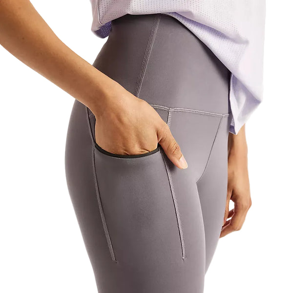 Der ses en person, der står med siden til, iført New Balance Reflective Rc Heat Legging 27″ i dameudgaven, farven Neptune Grey, med den ene hånd i sidelommen. Kun den nedre del af overkroppen og en del af den øvre del af kroppen er synlige, og tøjet er kombineret med en lys skjorte – hvilket gør dette outfit ideelt til løb i koldt vejr.