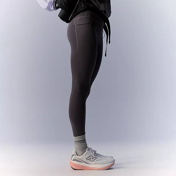 En person står i profil foran en ensfarvet lys baggrund iført New Balance Reflective Rc Heat Legging 27″ til kvinder i farven Neptune Grey, med synlige lommer og NB HEAT-teknologi. Outfitet er kombineret med lysegrå strømper og hvide løbesko med lyserøde såler – perfekt til løb i koldt vejr. Overkroppen er delvist uden for billedet.