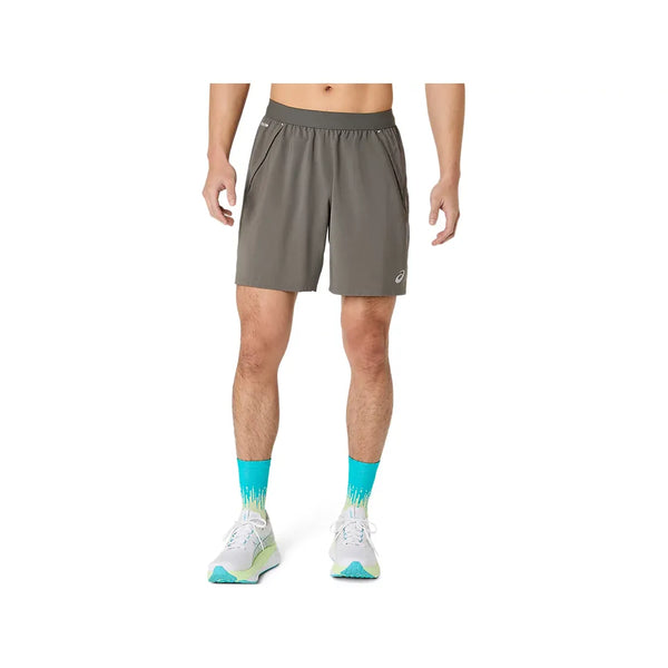 En person iført Asics Road 2-N-1 7In-shorts til mænd i farven Truffle Grey står foran en ensfarvet hvid baggrund. Fokus er på shortsene og underkroppen, hvor hænderne hviler afslappet langs siden, suppleret af lyseblå sokker og hvide løbesko.