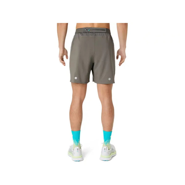 Der ses en person set bagfra, iført Asics Road 2-N-1 7In-shorts til mænd i farven Truffle Grey. Personen har desuden lyse, blågrønne mønstrede sokker og hvide løbesko på. Fokus er på personens lænd og ben foran en ensfarvet hvid baggrund.