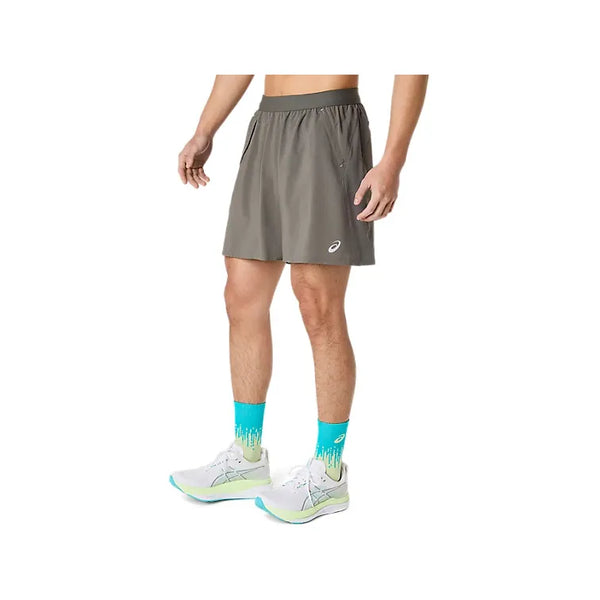 En person iført ASICS Road 2-N-1 7In-shorts til mænd i farven Truffle Grey, blågrønne sokker med grønne detaljer og hvide løbesko står foran en ensfarvet hvid baggrund, hvor man kan se vedkommende fra midten af overkroppen og ned til fødderne.