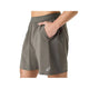 En person iført trøffelgrå ASICS Road 2-N-1 7In-shorts til mænd, med lommer og et ASICS-logo på venstre ben. Personens hånd er i den ene lomme, og billedet viser shortsene fra taljen til midt på låret.