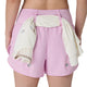 En person iført ASICS Road 3.5In Shorts Dame i farven Hazy Lilac med en hvid skjorte eller jakke, der er trukket gennem linningen bagpå, fotograferet bagfra foran en ensfarvet hvid baggrund.