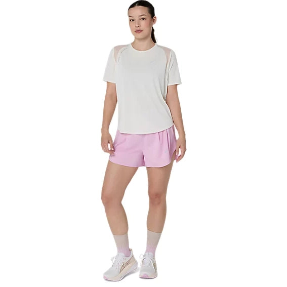 En kvinde står med ansigtet vendt fremad. Hun har en hvid kortærmet bluse på, Asics Road 3.5In Shorts Dame i farven Hazy Lilac, lyserøde sokker og hvide sneakers. Hendes hår er sat op, og armene hænger afslappet ned langs siden.
