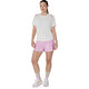 En kvinde står med ansigtet vendt fremad. Hun har en hvid kortærmet bluse på, Asics Road 3.5In Shorts Dame i farven Hazy Lilac, lyserøde sokker og hvide sneakers. Hendes hår er sat op, og armene hænger afslappet ned langs siden.
