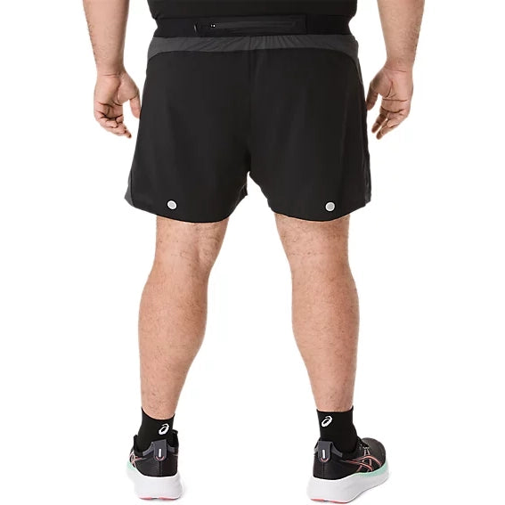 En person iført sorte Asics Road 5In-shorts, Herre i farvekombinationen Performance Black/Graphite Grey, samt sorte sokker og sorte løbesko med hvide såler, står med ryggen til kameraet. Den ensfarvede hvide baggrund fremhæver dette klassiske løbetøj fra Asics.