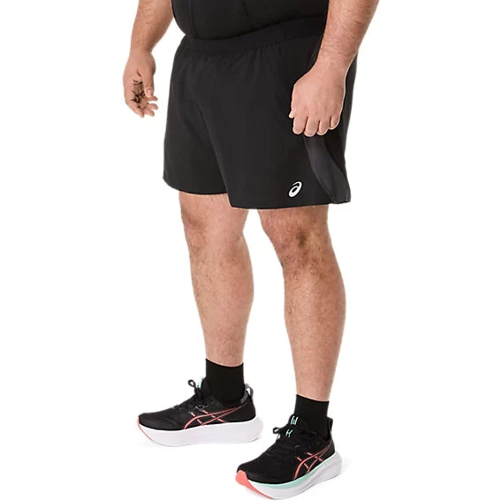 En person står iført et par ASICS Road 5In Shorts, Herre, i farvekombinationen Performance Black/Graphite Grey, sammen med sorte ankelstrømper og sorte løbesko med hvide såler og lyserøde detaljer. Billedet er beskåret ved overkroppen og placeret på en ensfarvet hvid baggrund, hvilket fremhæver det sportslige løbelook.