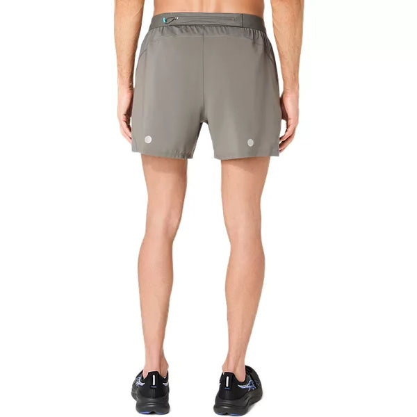 En person står med ryggen til kameraet iført Asics Road 5In Shorts, Herre, i farven Truffle Grey samt sorte løbesko foran en ensfarvet hvid baggrund.