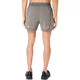 En person står med ryggen til kameraet iført Asics Road 5In Shorts, Herre, i farven Truffle Grey samt sorte løbesko foran en ensfarvet hvid baggrund.