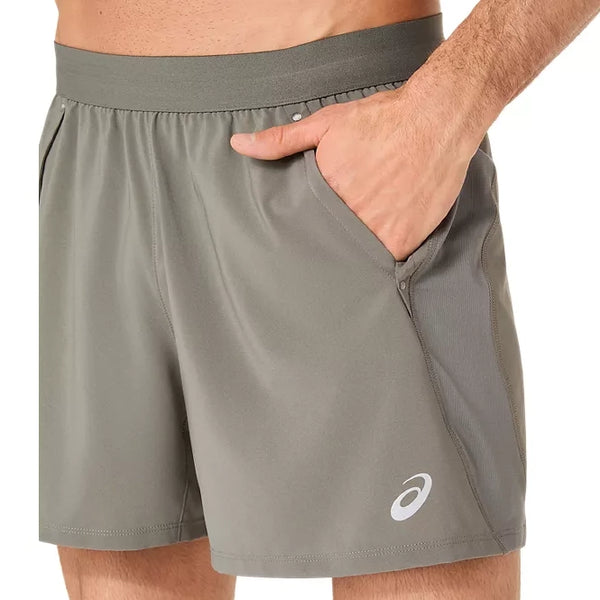 En person har Asics Road 5In Shorts, Herre i farven Truffle Grey på, med elastisk linning og sidelommer. Det hurtigtørrende stof og det synlige Asics-logo tæt på nederkanten sikrer komfort og ydeevne. Den ene hånd hviler i en lomme.