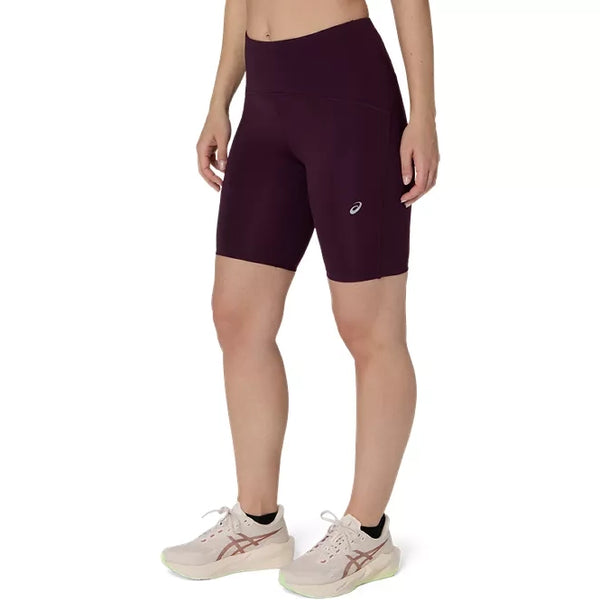 En person har Asics Road High Waist 8In Sprinter-shorts i farven Dark Aubergine og lyserøde sportssko på, og billedet viser vedkommende fra taljen og ned mod en hvid baggrund.