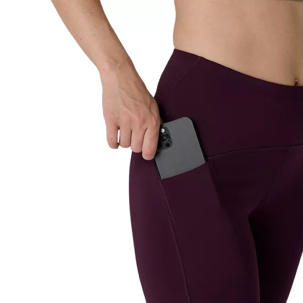 En kvinde iført Asics Road High Waist 8In Sprinter løbetights, Dame, Dark Aubergine lægger en sort smartphone i sidelommen. Kun hendes overkrop og øvre lår er synlige.
