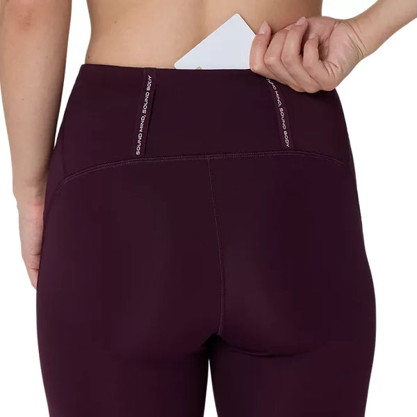 En person lægger et hvidt, rektangulært objekt ned i lommen i linningen på et par mørke, auberginefarvede Asics Road High Waist 8In Sprinter-tights til kvinder. Kun vedkommendes overkrop og hånd er synlige.