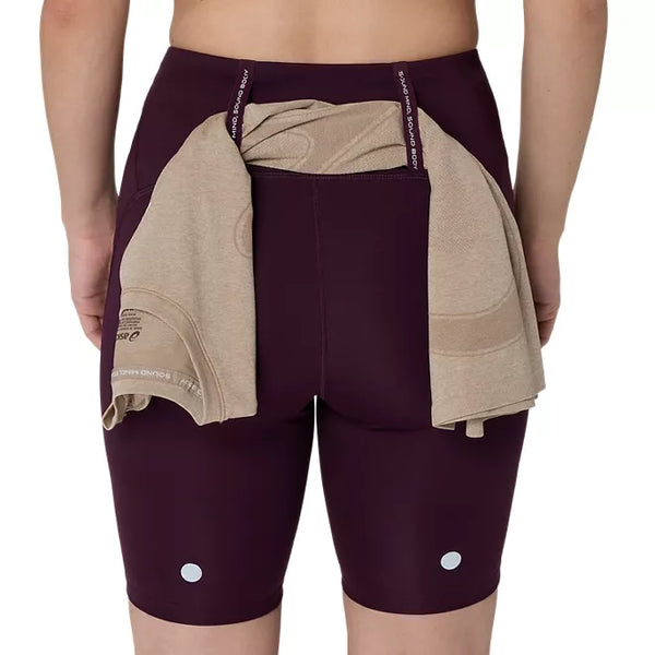 Der ses en person set bagfra iført ASICS Road High Waist 8In Sprinter-shorts til kvinder i farven Dark Aubergine. En beige skjorte er stukket ned i linningen, og der ses to reflekterende prikker på den nederste del af løbetights til kvinder.