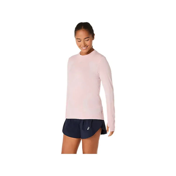 En kvinde iført en Asics Road Seamless LS Top i Morganite/Dusty Red og marineblå sportsshorts står på en almindelig hvid baggrund og kigger ned med et lille smil.