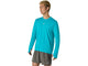 En person med kort, lyst hår har ASICS Road Seamless LS Top, Herre, i farven Energy Aqua på – en åndbar langærmet trøje – kombineret med grå shorts, hvor begge dele er forsynet med ASICS-logoet, mens vedkommende står foran en ensfarvet hvid baggrund.