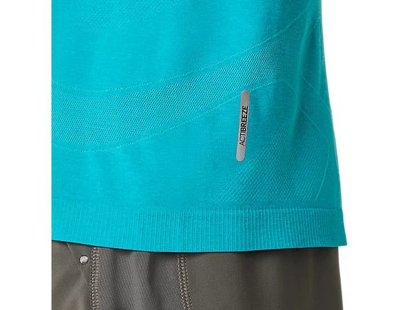 Et nærbillede af den nederste del af en blågrøn Asics Road Seamless Ls Top, Herre, Energy Aqua, hvor der er et lille reflekterende mærke med teksten ADIBREEZE, kombineret med grå sportsshorts med synlige sømme. Designet, der minder om en åndbar langærmet trøje, lægger vægt på åndbarhed og komfort, hvor kun overkroppen og en del af shortsene er synlige.