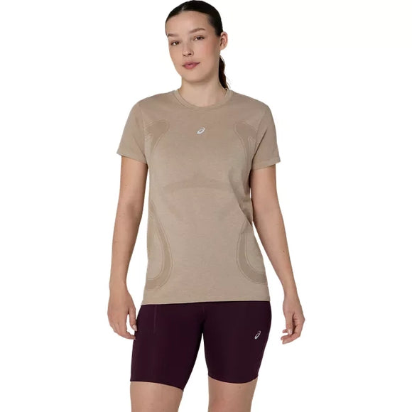 En person står med ansigtet vendt fremad iført en Asics Road Seamless SS Top, Dame, i farven greige med et lille logo på brystet og mørkeburgunderfarvede ROAD-træningsshorts foran en ensfarvet hvid baggrund.