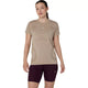 En person står med ansigtet vendt fremad iført en Asics Road Seamless SS Top, Dame, i farven greige med et lille logo på brystet og mørkeburgunderfarvede ROAD-træningsshorts foran en ensfarvet hvid baggrund.