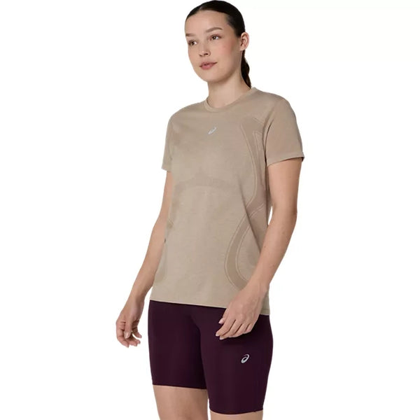 En kvinde står let vendt væk fra kameraet iført en Asics Road Seamless SS Top i farven »greige« og maroon-farvede shorts foran en ensfarvet hvid baggrund.