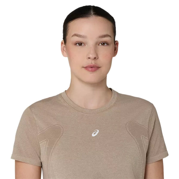 En person med glat, mørkt hår, der er sat op, har en Asics Road Seamless Ss Top, Dame, Greige, på og ser ind i kameraet med et neutralt udtryk foran en ensfarvet hvid baggrund.