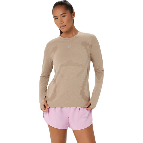 En person står med ansigtet vendt fremad iført en gråbeige Asics Road Seamless SS Top, Dame, og lyserøde sportsshorts foran en ensfarvet hvid baggrund.