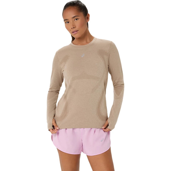 En person står med ansigtet vendt fremad iført en gråbeige Asics Road Seamless SS Top, Dame, og lyserøde sportsshorts foran en ensfarvet hvid baggrund.