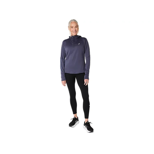 En person står med ansigtet vendt fremad og smiler, med lyst hår sat op, iført en mørk langærmet sportstrøje, sorte Asics Road Seamless Tight (dame) i farven Performance Black og sorte løbesko. Baggrunden er helt hvid.