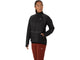 En person iført en Asics Road Winter Run-jakke til kvinder i farven Performance Black, med lynlås og lommer, kombineret med bordeauxrøde leggings, står foran en ensfarvet hvid baggrund og kigger til siden.