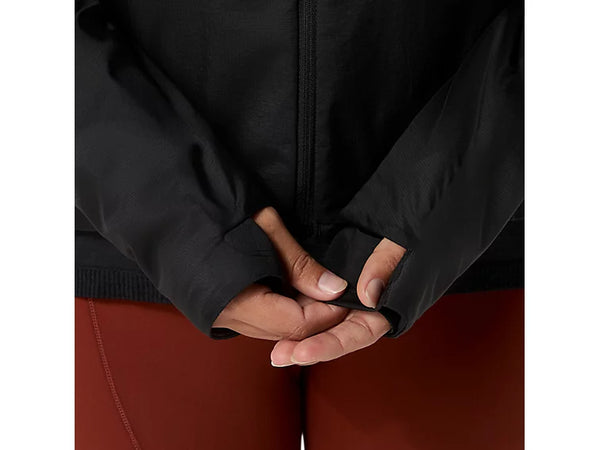 En person står med hænderne samlet og er iført ASICS Road Winter Run Jacket, Dame i farven Performance Black – en vinterløbejakke med tommelfingerhuller – samt røde bukser. Fokus er rettet mod jakkens ærmer og hænderne, hvilket fremhæver det gennemtænkte design af denne løbejakke til kvinder fra ASICS, mens baggrunden forbliver ude af billedet.
