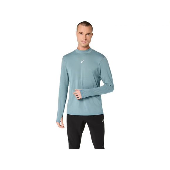 En mand står foran en hvid baggrund iført Asics Road Winter Seamless Ls Top til mænd i farven »Foggy Teal«, der er fremstillet af termisk materiale med tommelfingerhuller i ærmerne og et lille logo på brystet. Trøjen er designet som en sømløs vinterløbetrøje og giver optimal komfort i koldt vejr. Han har den på sammen med sorte bukser.