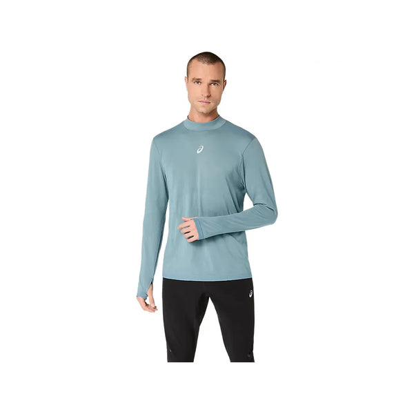 En mand står foran en hvid baggrund iført Asics Road Winter Seamless Ls Top til mænd i farven »Foggy Teal«, der er fremstillet af termisk materiale med tommelfingerhuller i ærmerne og et lille logo på brystet. Trøjen er designet som en sømløs vinterløbetrøje og giver optimal komfort i koldt vejr. Han har den på sammen med sorte bukser.