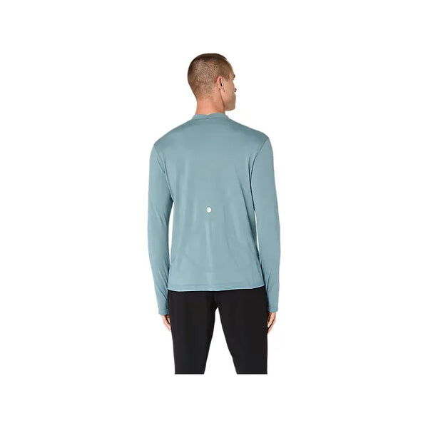 En person står med ryggen til og har en Asics Road Winter Seamless LS Top til mænd i farven »Foggy Teal« på samt sorte bukser. Denne langærmede trøje, der er fremstillet af termomateriale, har en lille hvid prik øverst på ryggen. Baggrunden er ensfarvet hvid.