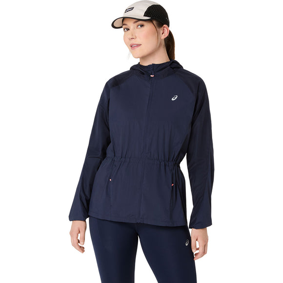 En kvinde iført Asics Road Packable Jacket, Dame, i Midnight og matchende leggings står med den ene hånd ved siden af sig. Jakken, der er lavet af genbrugsmaterialer, har et lille hvidt logo på brystet. Hun bærer også en sort og hvid kasket.