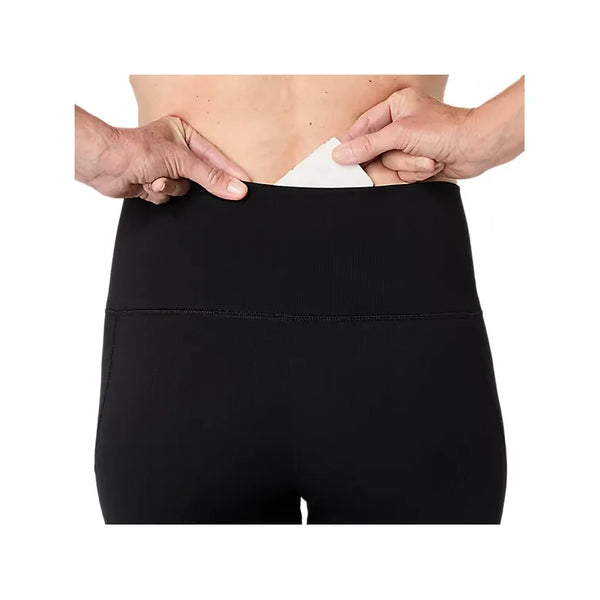 En person stikker en hvid, rektangulær genstand – muligvis et kort eller et stykke papir – ind i lommen i linningen på et par sorte Asics Road Seamless Tight-leggings til kvinder (Dame) i farven Performance Black. Billedet fokuserer på den nedre del af ryggen og hænderne og fremhæver det praktiske design af disse Asics-sportstights.