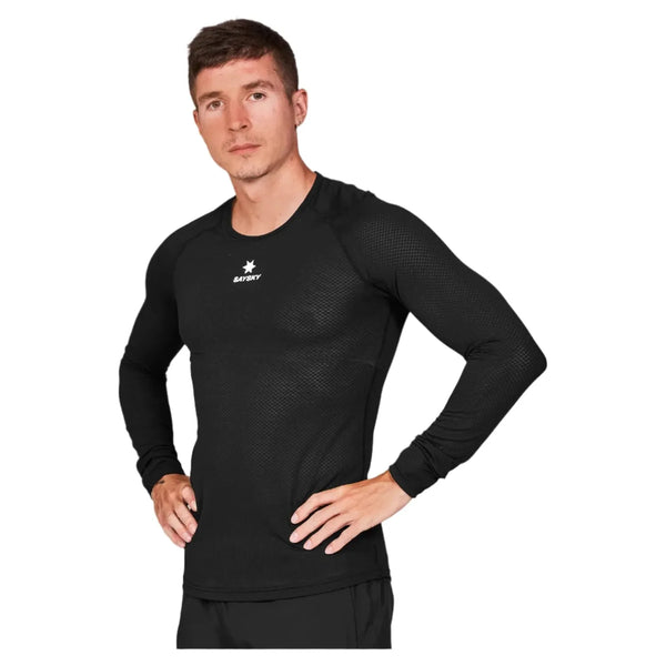 En person står med hænderne på hofterne i en tætsiddende sort Saysky Mesh Base Layer LS løbetrøje, Unisex, med en lille hvid stjerne og "SAFARY" på brystet, lavet af svedtransporterende mesh-stof, mod en almindelig hvid baggrund.