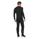 En mand kigger væk, iført Saysky Mesh Base Layer LS i sort og matchende bukser med hvid tekst på højre ben, sammen med sorte sportssko. Dette svedtransporterende unisex-outfit skiller sig ud mod en almindelig hvid baggrund.