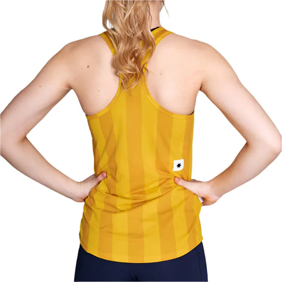 En kvinde med langt, bølget blondt hår ses bagfra iført Saysky W Stripe Combat Singlet, Dame, Yellow Aop og mørke bukser. Hun står med hænderne på hofterne mod en hvid baggrund - perfekt som en fugttransporterende løbetop.