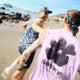 To mennesker løber langs en solrig sandstrand ved vandet. Den ene har en blomstret tank på, den anden har Saysky Flower Flow T-shirt, Herre, Purple - og viser sommerstil og komfort i en energisk scene under blå himmel.