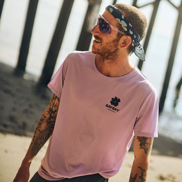 En tatoveret mand i en lilla Saysky Flower Flow T-shirt, mønstret pandebånd og solbriller står smilende på en strand med træmoler i baggrunden.
