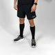 En person står på en hvid baggrund iført en sort T-shirt, sorte shorts med Lifeguard-logo, LiiteGuard Shin-Tech Light Running Socks (Unisex, Black/Grey), sorte og hvide sportssko og et sort smartwatch. Deres ansigt er ikke synligt.