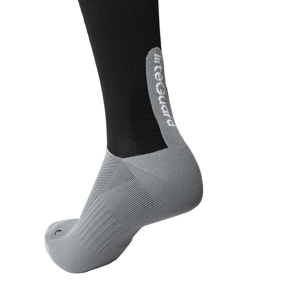 Et nærbillede af en persons underben iført Liiteguards Shin-Tech Light Running Sock, Unisex, i sort/grå. Foden er grå med rillede detaljer, og på hælen står mærkets navn i hvidt på et åndbart materiale.