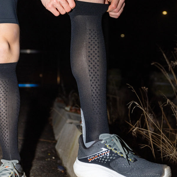 Om natten justerer en person udendørs en LiiteGuard Shin-Tech Light løbestrømpe (Unisex, sort/grå) med tørt græs og fortov i baggrunden. Personen har også grå Saucony-løbesko på.
