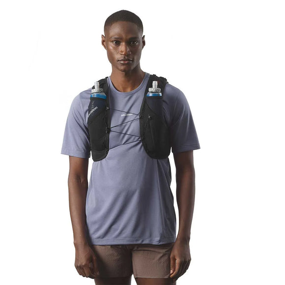 En person i en lyslilla kortærmet skjorte, brune shorts og en sort Salomon ADV Skin 5 Løbevest, Unisex (med to synlige vandflasker) står på en almindelig hvid baggrund.