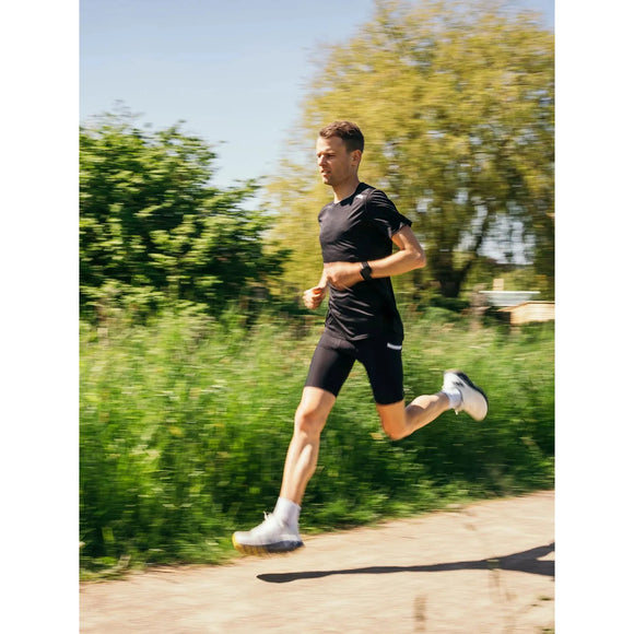 En mand iført Fusion's Short Tights Advanced, Unisex, i Superblack, sammen med hvide sko, løber udenfor på en asfalteret sti ved siden af grønt græs og træer på en solrig dag.