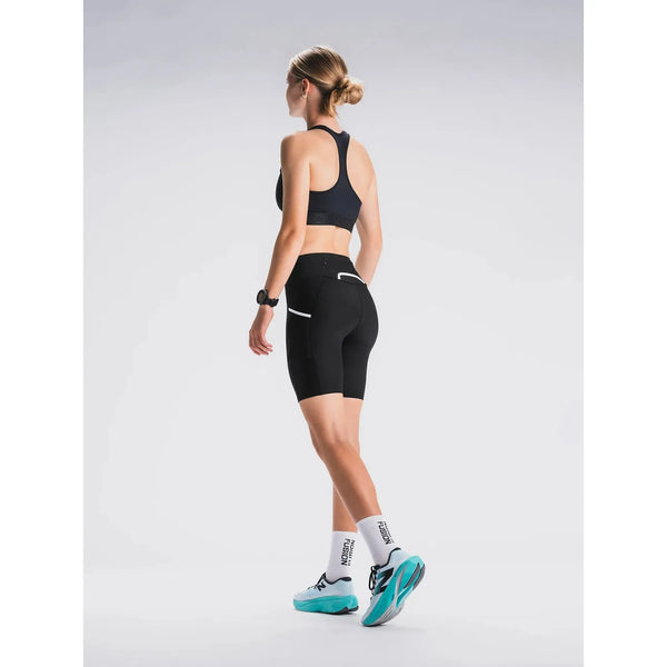 En person med lys hud og tilbagestrøget blondt hår er iført sort sports-bh, Fusion Short Tights Advanced Unisex i Superblack, hvide sokker og blågrønne løbesko - perfekt til løb eller træning - og står og kigger væk på en almindelig lys baggrund.